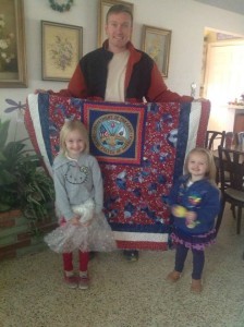 Two children pose with a woman holding a colorful quilt indoors.