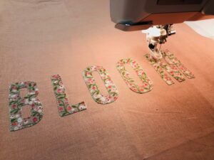 Sewing machine stitching the word 'BLOOM' made of floral fabric.