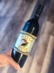 A hand holds a bottle of Witch Wine with a witch illustration on the label.